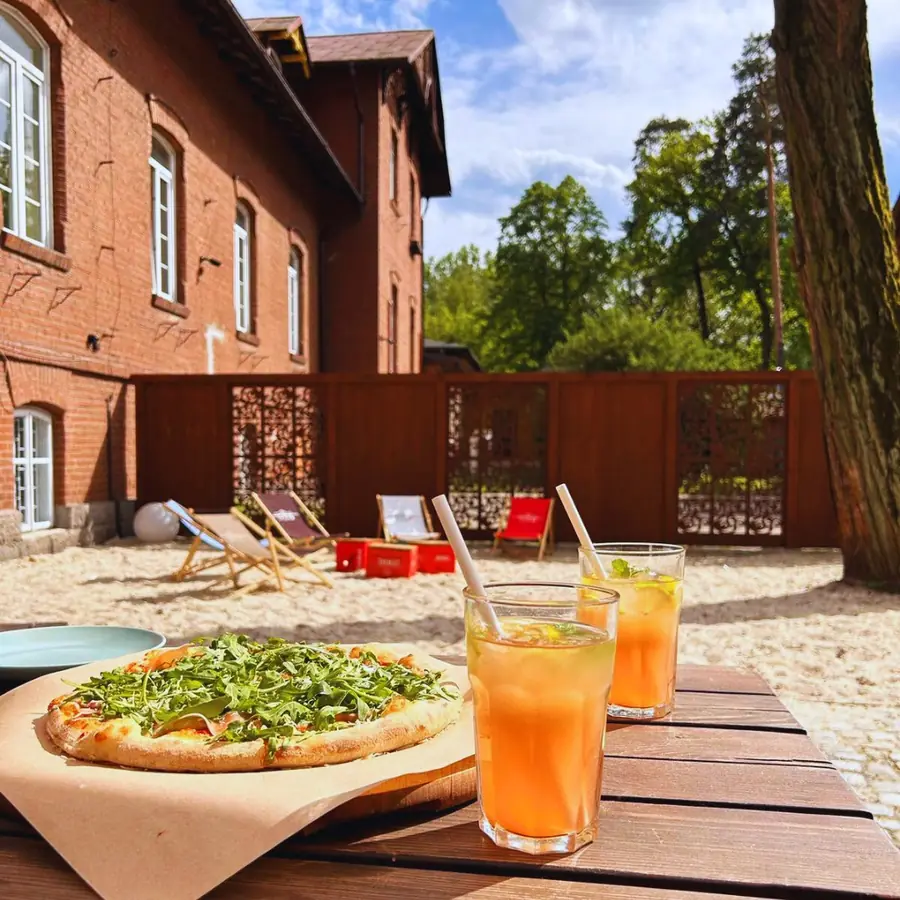 Ogródek przy ceglanym budynku: pizza i dwa mrożone napoje na drewnianym stole.
