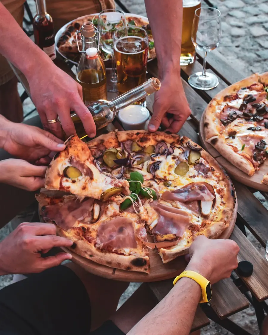 Kilka par rąk sięga po kawałki pizzy nad stołem zastawionym butelkami.