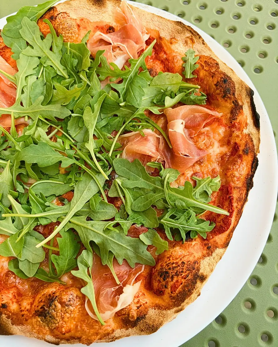 Pizza z prosciutto i rukolą na białym talerzu.